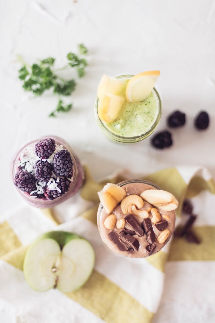 lemons and blackberries, cashews and banana slices, little milk chocolate bits, topping three different glasses, filled with blended drinks, protein shake recipes, halved green apple nearby