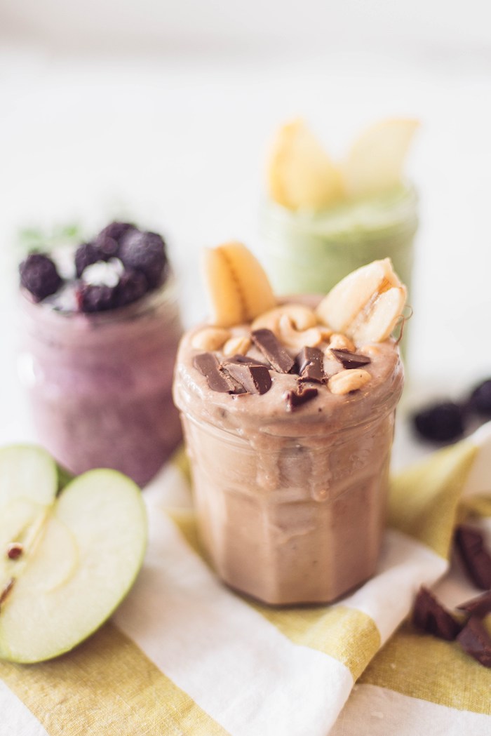 half of a green apple, near glass containing a beige smoothie, topped with banana slices, cashews and chocolate bits, protein shake recipes, more glasses in the background