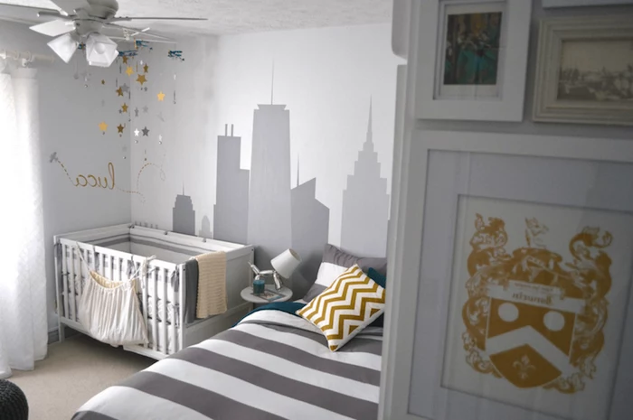bed with striped duvet, in dark gray and white, near white and gray baby crib, wall mural depicting a city scape, painted in gray, various yellow motifs