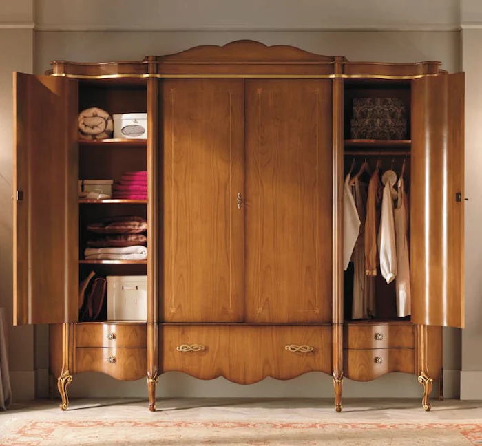 brown closet with four doors, made from wood, with engraved details, and ornate handles, two doors are open, to reveal clothes, storage boxes and blankets, classical wardrobes