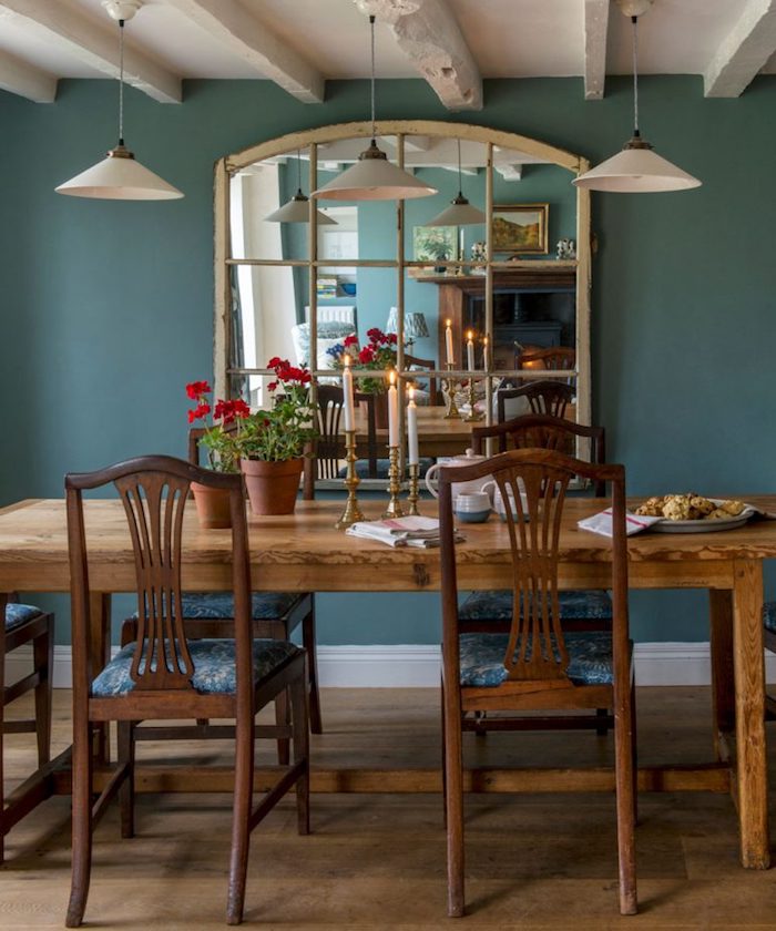 teal wall with large mirror, near wooden dining table, country kitchen décor, with three candles, flowerpots and crockery, white ceiling with wooden beams