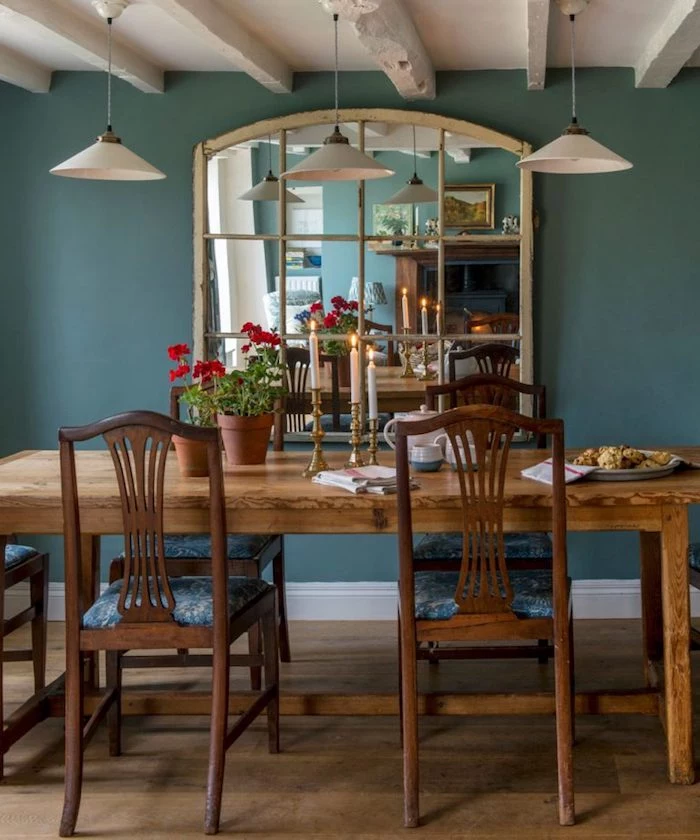 teal wall with large mirror, near wooden dining table, country kitchen décor, with three candles, flowerpots and crockery, white ceiling with wooden beams