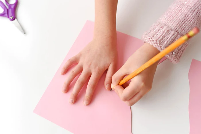 child in pale pink knitted sweater, tracing her hand with pencil, on sheet of pale pink paper, easter diy, scissors nearby