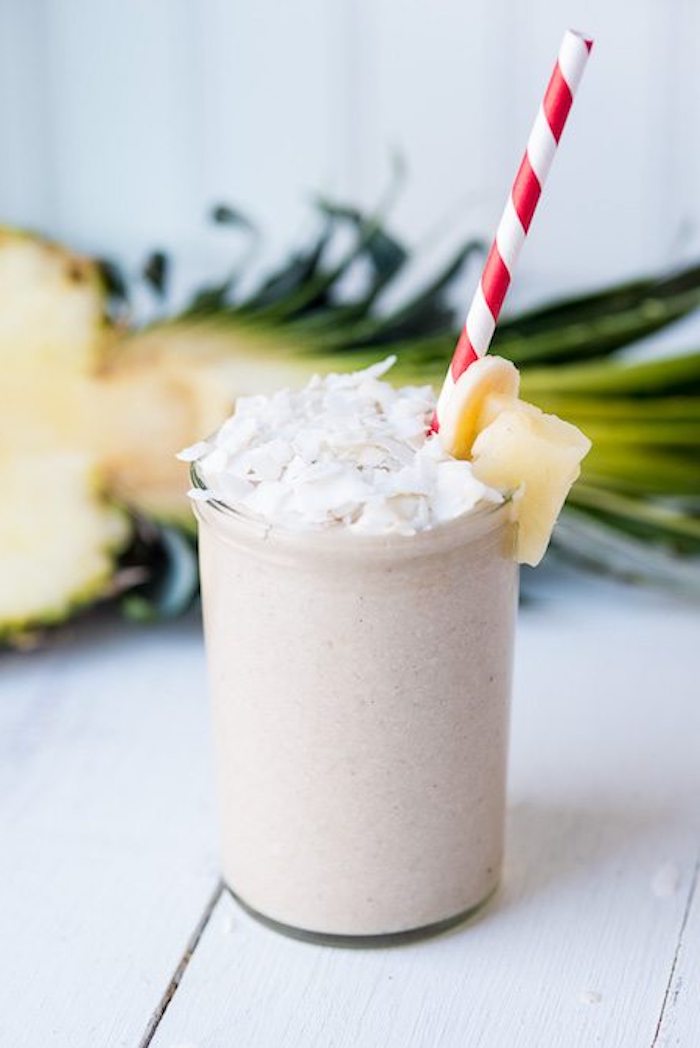 plain clear glass, filled with beige creamy drink, topped with coconut flakes, banana and pineapple chunks, fruit smoothie recipes, halved pineapple in the background