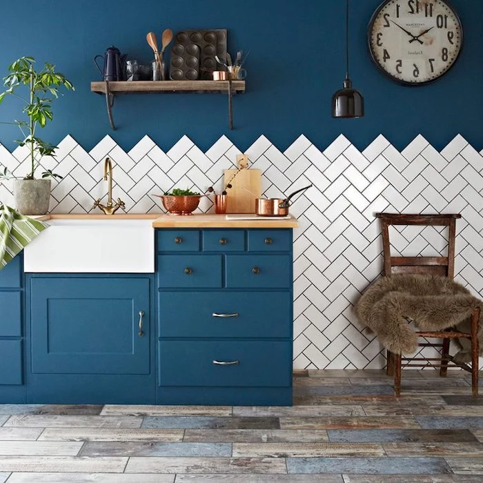 blue vintage rustic kitchen cabinets, in room with grayish brown laminate floor, dark blue wall half covered in white tiles, antique chair and wall closk