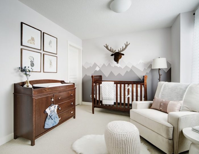 neutral colors in nursery room, dark wooden crib and matching changing table, four framed images, wall mural with gray mountains, pale white armchair