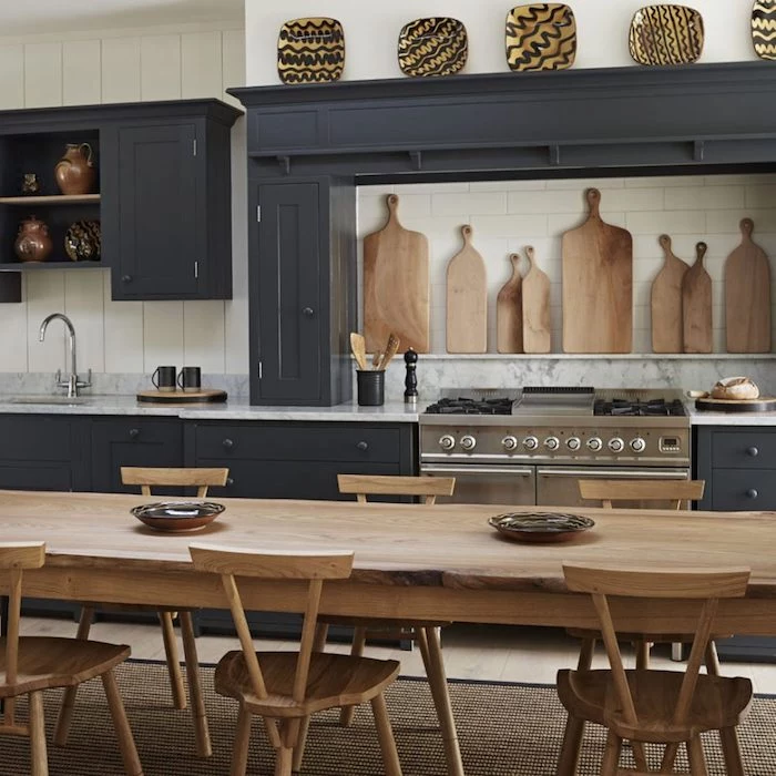 large wooden dining table, with six matching chairs, in room with dark gray, rustic kitchen cabinets, decorated with hand-painted wooden plates, and cutting boards