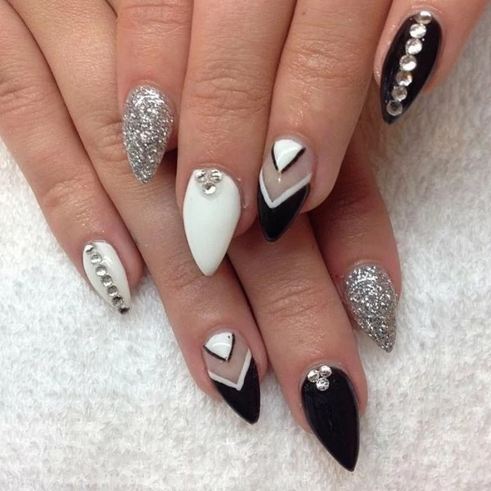 triangular shapes on short stiletto nails, painted in black and white nail polish, decorated with silver glitter, and round rhinestone stickers