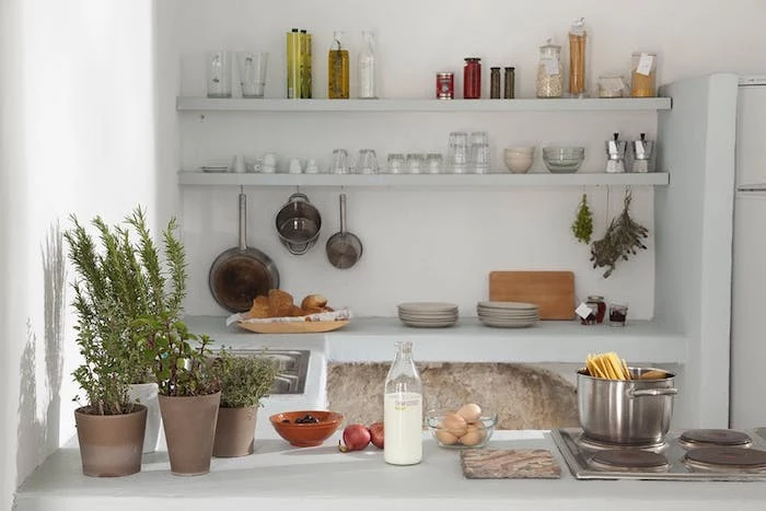 rustic kitchen in white, pale gray shelves, with cups and glasses, pans and condiments, several potted plants, milk and eggs, cooking dish with pasta