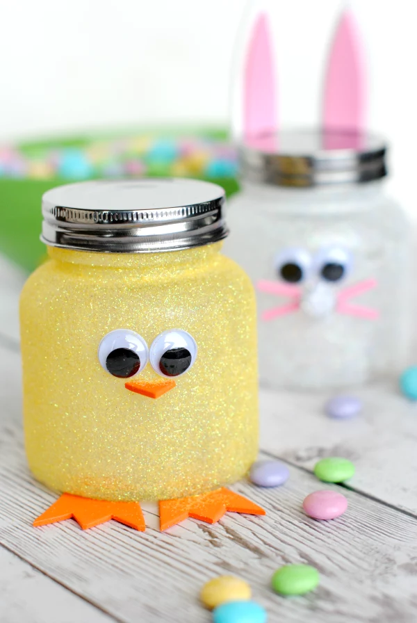 jars with metal lids, covered with yellow and white glitter, and decorated with felt cutouts, and eye stickers, to look like easter chick and bunny, easter crafts for adults, colorful candy nearby