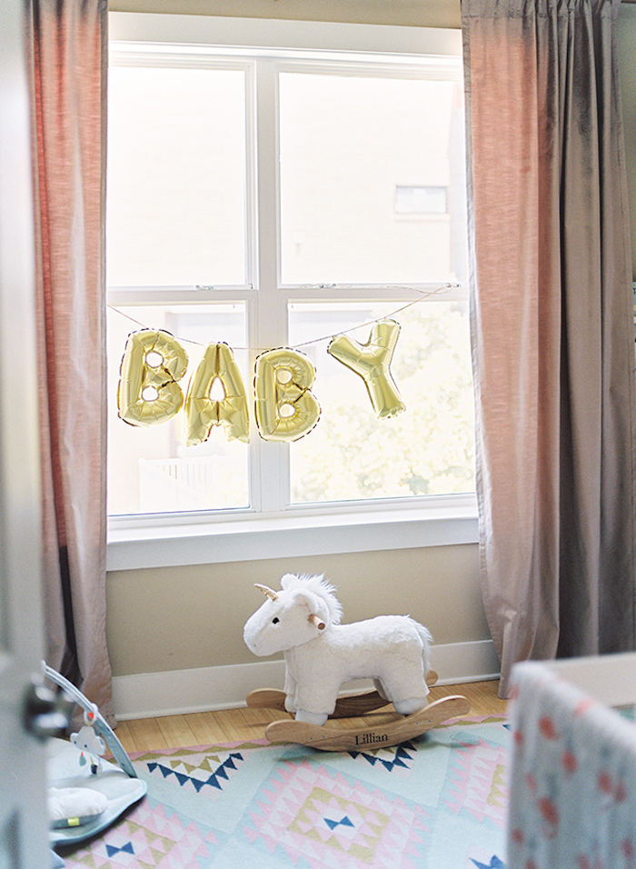 baloons in shiny golden color, shaped like letters, spelling the word baby, hung on a window, with pastel pink curtains, baby girl themes, rocking unicorn toy