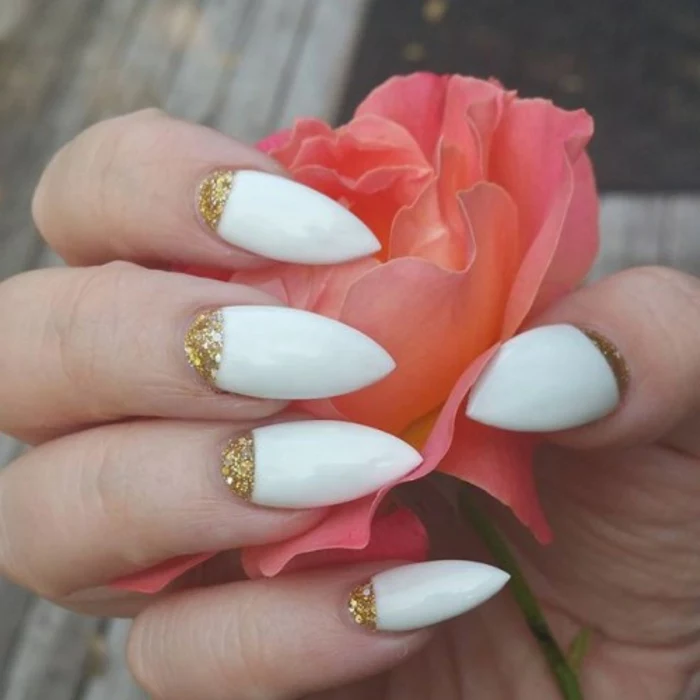 hand with long sharp nails, stilleto nail designs, painted in white nail polish, and decorated with golden glitter, holding a faux pink flower