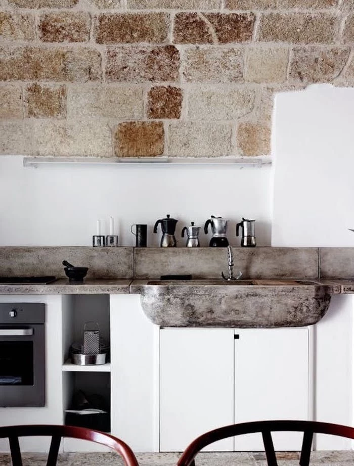 stone sink basin, and antique metal tap, in rustic kitchen, with white cupboards, and white wall with beige stone brick details, small assortment of coffee pots
