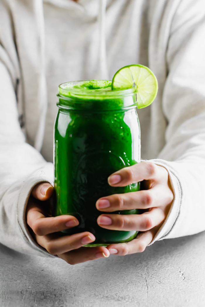 woman in pale grey hoodie, holding a jar, filled with a bright green smoothie, and decorated with a lime slice