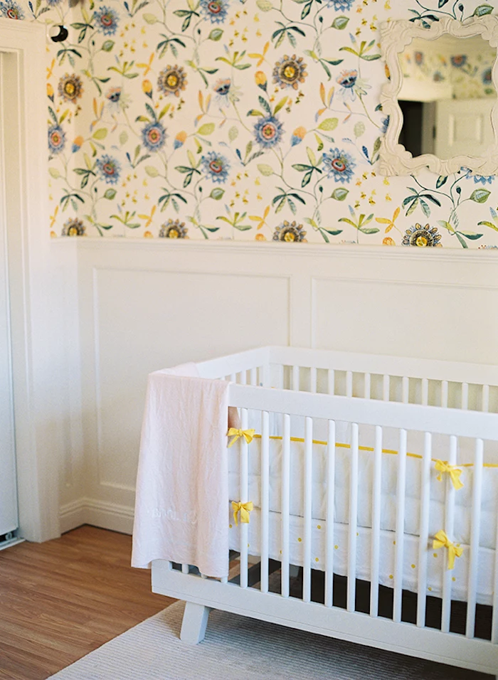 paneling in white, and white wallpaper, with flower pattern in yellow, blue and green, near white crib with yellow bows, decorative mirror in ornate frame