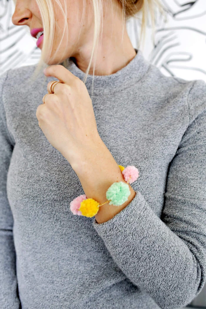 blonde woman with tied back hair, wearing gray jumper, and cyclamen pink lipstick, with bracelet made of pompoms, gift ideas for mom