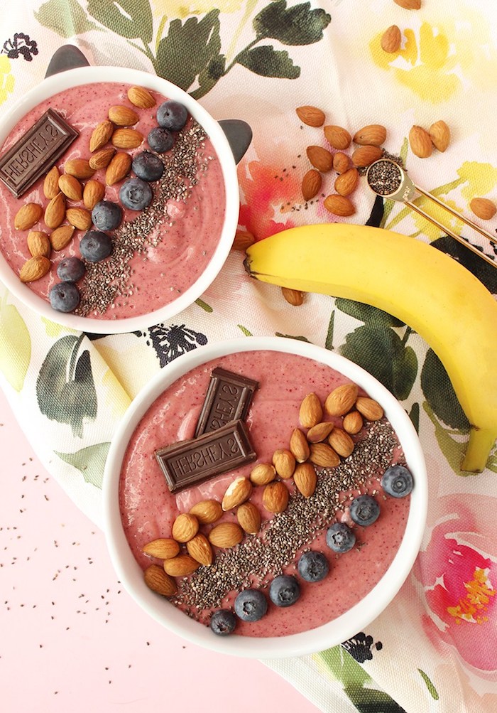raw almonds and blueberries, small chocolate blocks and chia seeds, topping two bowls, filled with pink berry puree, healthy breakfast smoothies, banana nearby