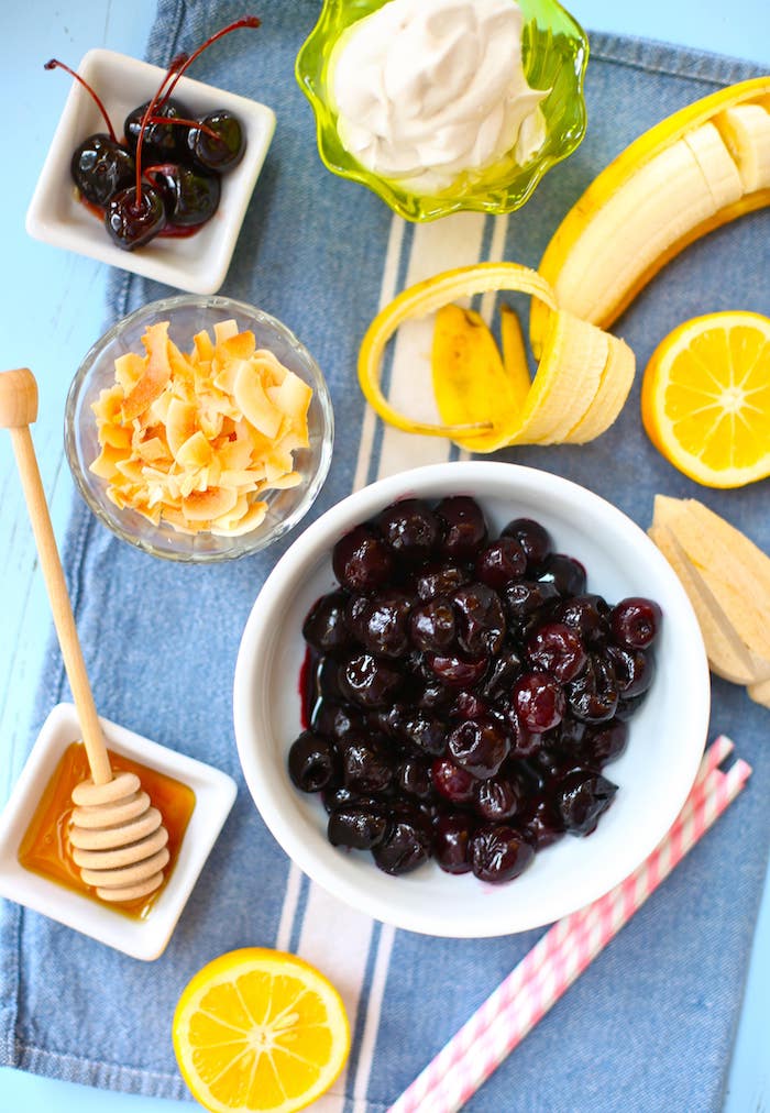 white bowl of maraschino cherries, placed on a table cloth, near cup of yoghurt, little plate with honey, peeled banana and cut lemon, toasted coconut flakes, healthy smoothie recipes