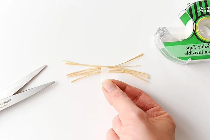 hand sticking a bunch of straw-like whiskers together, using white sticky tape, craft ideas for kids, sticky tape dispenser, and scissors nearby
