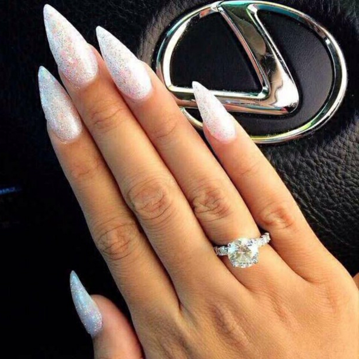 diamond ring on hand with long, slender fingers and sharp, stiletto acrylic nails, covered in iridescent silver glitter, lexus logo in background