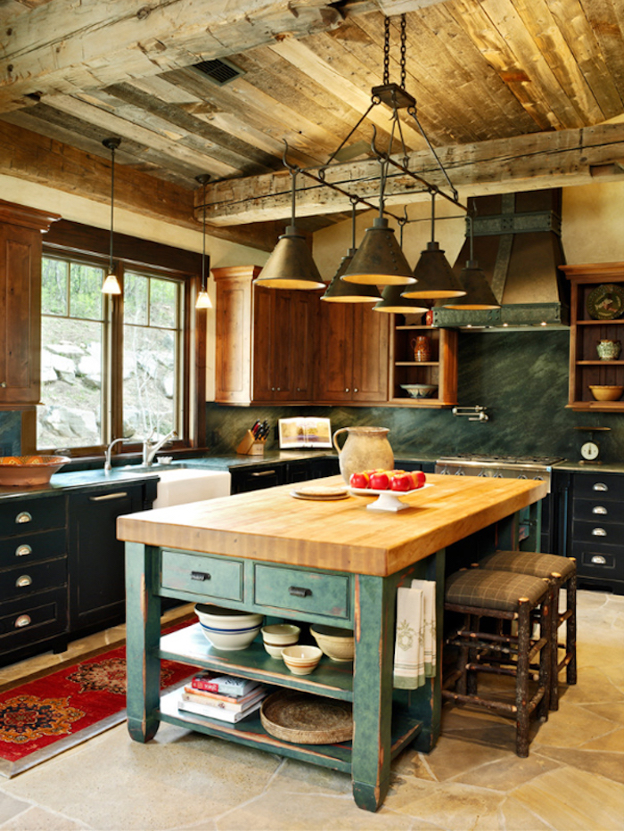 brown and black country kitchen cabinets, and a shabby chic wooden kitchen island, with two hand-made stools, inside room with wooden beams, and beige stone floor