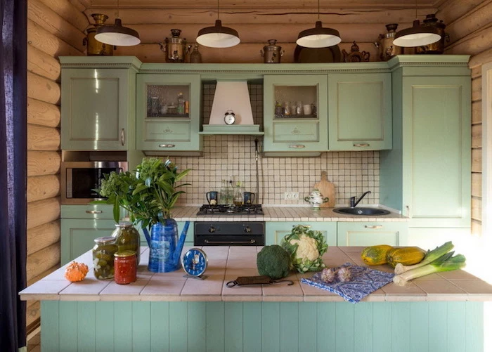 minty green country kitchen cabinets, with a matching kitchen island, vegetables and canned preserves in jars, four hanging lamps, walls covered with wooden planks