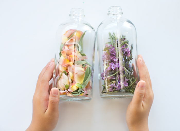 child's hands holding two clear bottles, made of plastic or glass, filled with rose petals and sage, lilac and rosemary, mother's day gift ideas, home-made perfume