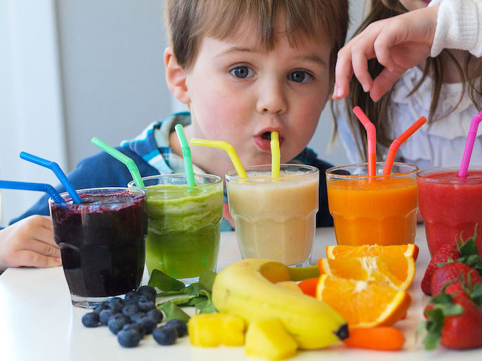 toddler drinking juice from a straw, near five clear glasses, each containing a juice in a different color, banana and orange, berries and carrots, spinach smoothie 