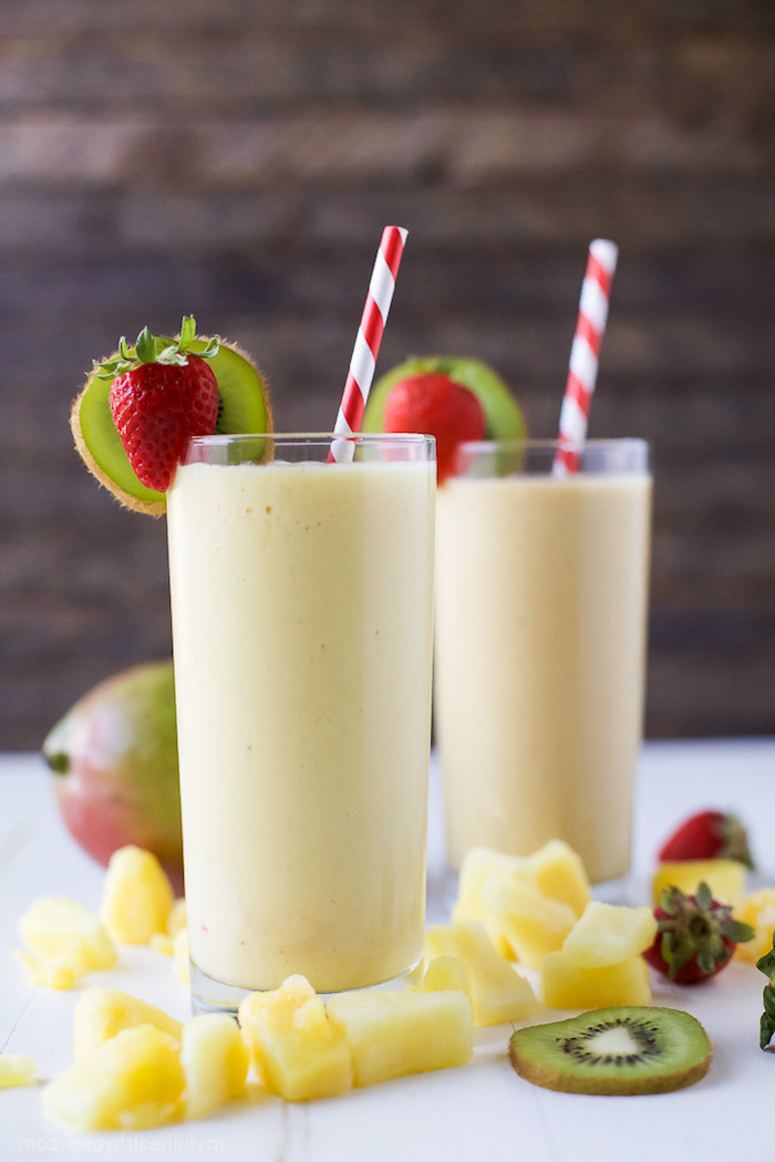 mango chunks and kiwi slices, strawberries and a whole mango, near two glasses, decorated with kiwi and strawberries, filled with creamy yellow liquid, smoothie recipes