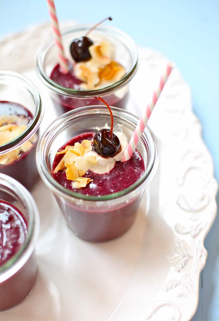 close up of four small jars, containing burgundy red mix, healthy smoothie recipes, topped with toasted coconut flakes, cream and a maraschino cherry
