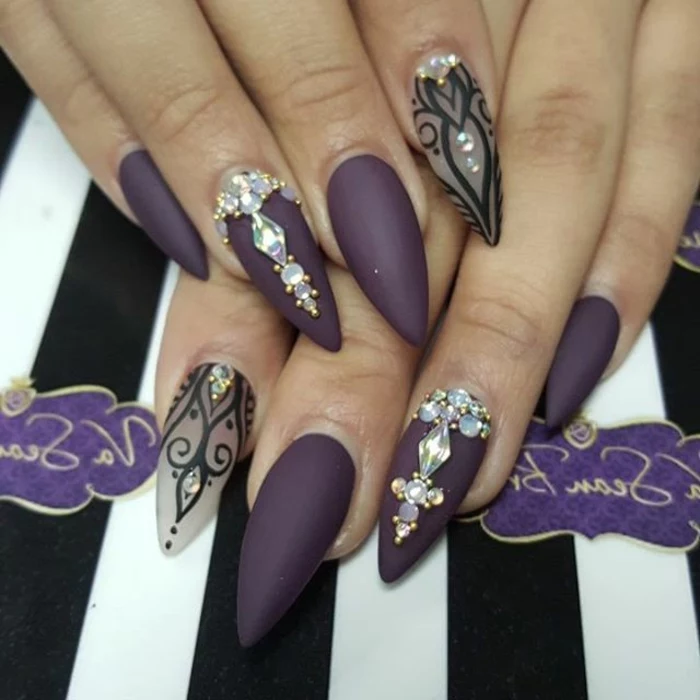 long stiletto nails, painted in matte purple, and clear nail polish, with black hand-drawn details, and iridescent nail decal gems