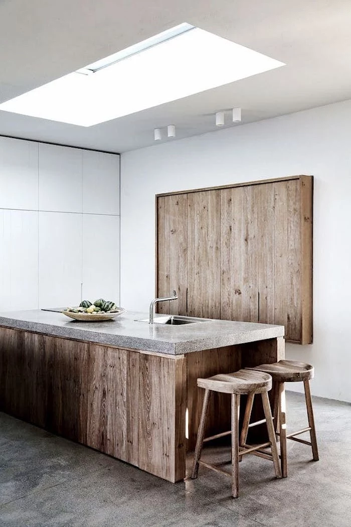 large kitchen island made of wood, with marble-like top surface, featuring an inbuilt metal sink, and a plate with fruit, two stools and a decorative wooden board nearby, rustic country home décor, white walls and minimalist aesthetic