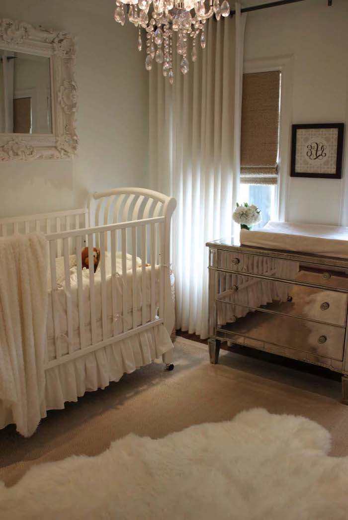 baroque framed mirror, white crystal chandelier, near mirrored chest of drawers, baby nursery with white crib, wooden floor with white lambskin rug