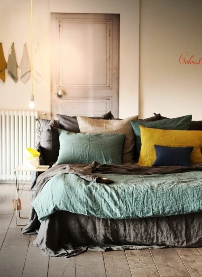 pillows in light teal and brown, dark blue yellow, on bed with matching covers, in room with light brown wooden floors, accent color with gray walls