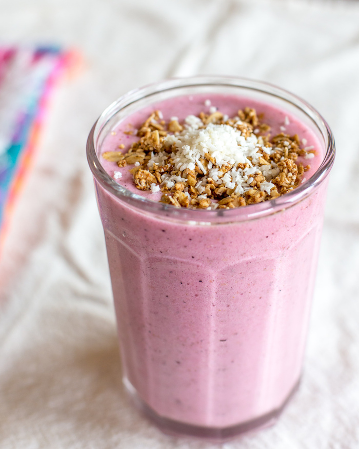 smoothie recipes, crushed beige nuts, and fine coconut flakes, topping a pale pink blended drink, inside a clear glass