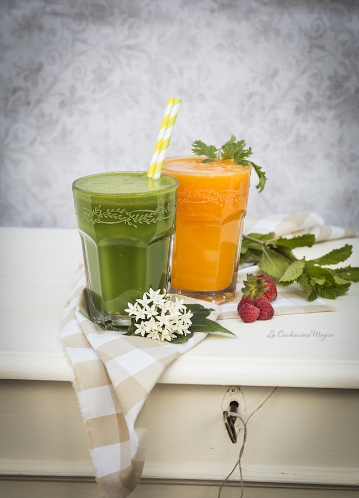 orange and green blended juice, in two clear decorated glasses, green smoothie recipe, placed on a chequered cloth, near orange blossoms, berries and mint
