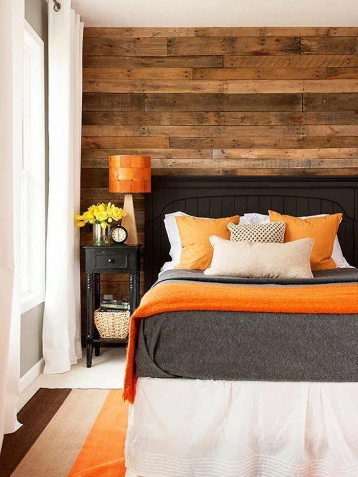 warm wooden planks, on bedroom wall, opposite wall is gray, dark grey bed, with light gray and beige, and orange bedcovers and pillows, colors that go with gray walls, striped three tonal rug