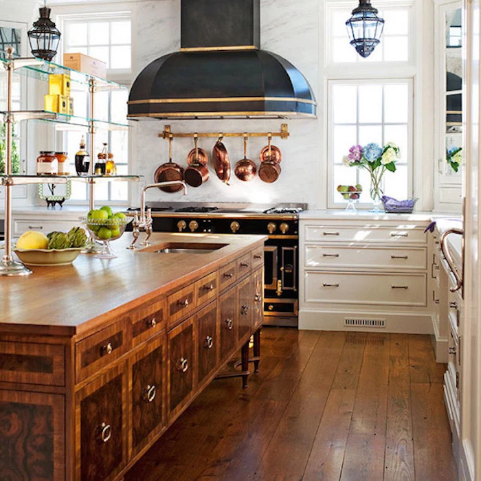 brass pots and pans, hanging over an antique black stove, with gold-colored details, near wooden country kitchen cabinets, in white and brown