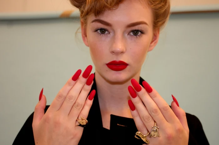 blonde woman with hair tied up, dressed in black, and holding up her hands, with long oval red manicure, and lipstick in matching color