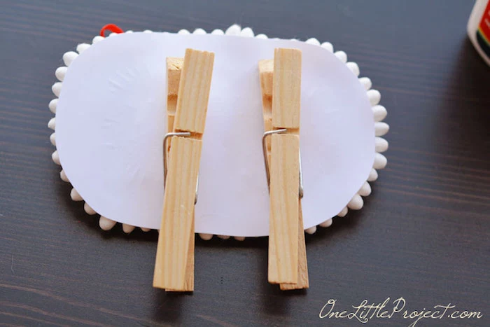 two plain wooden clothespins, stuck on the back of the lamb's paper body, craft ideas for kids, dark wooden surface 