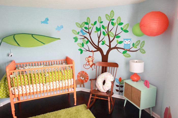 decal with a cartoon tree, birds and animals, on pale blue wall, near light turquoise cupboard, baby girl room décor, orange crib with light green bedding, and vintage rocking chair