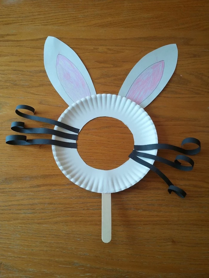 simple rabbit mask, made from white paper plate, with round hole, curled black paper whiskers, easter crafts for preschoolers, and hand-decorated ears, wooden ice cream stick holder