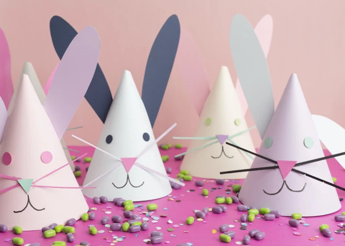 several paper party hats, in pale pastel colors, hand-made and decorated to look like bunnies, on pink surface, with green and purple candies