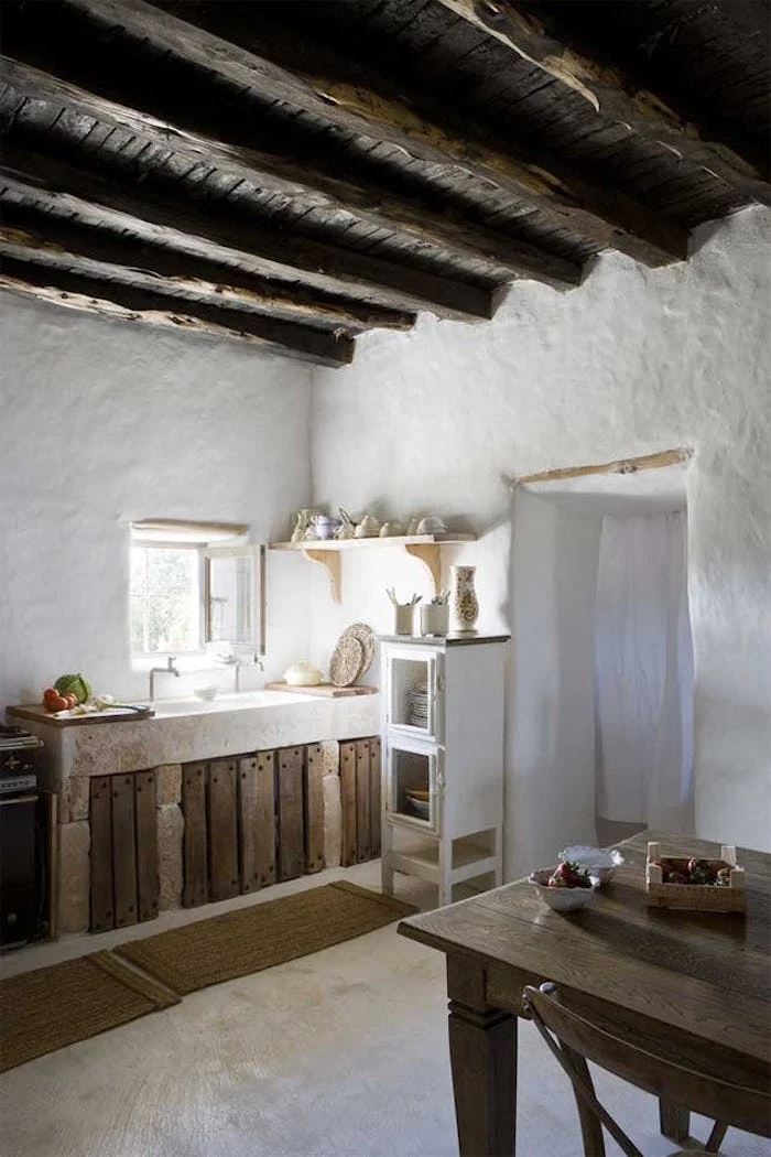 room with asymmetrical walls, covered in lime plaster, dark wooden ceiling with several large beams, country kitchen décor, antique sink near a small open window, dining table and chair