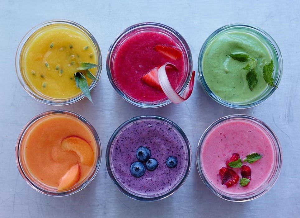 six identical glasses, each containing a different blended fruit drink, yellow and green smoothie, red and orange drink, purple and pink beverage 