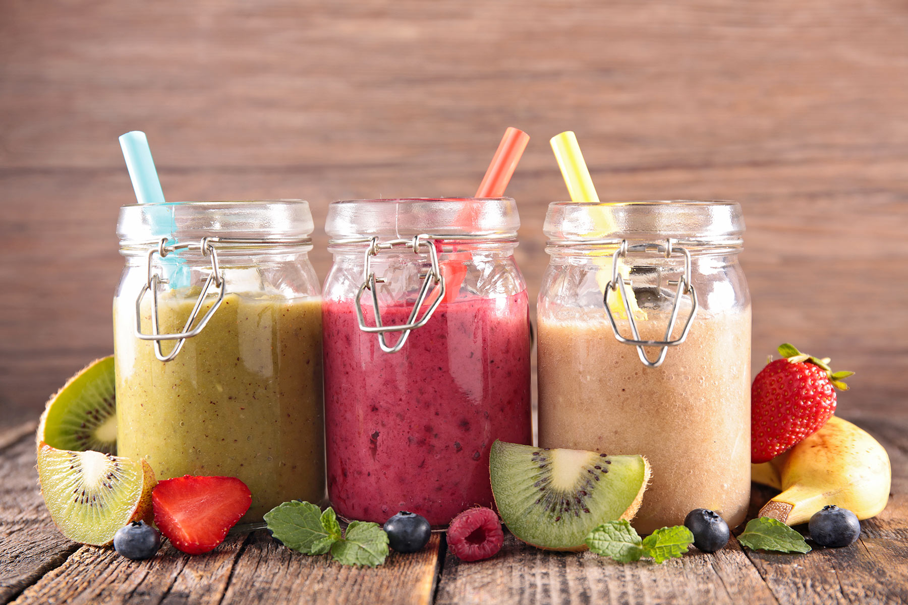 slices of kiwi and strawberry, blueberries and mint, near three jars with snap lids, containing blended drinks in different colors, healthy smoothie recipes, straws and a banana