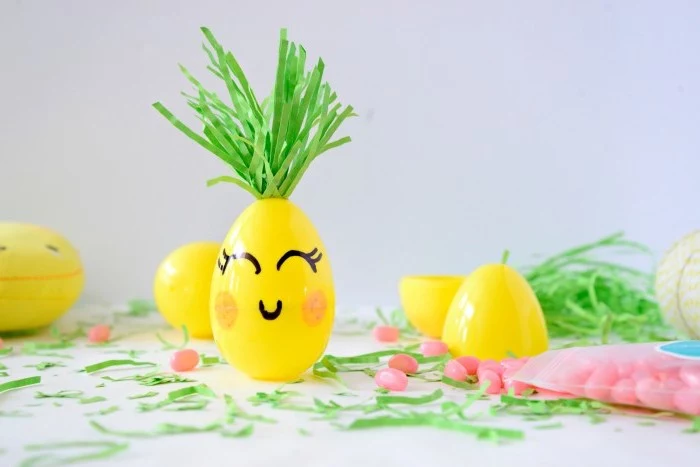 plastic yellow easter egg, decorated to look like a pineapple, with green felt leaves, and smiling face, drawn in marker, easter grass and candies, and other plastic yellow eggs nearby