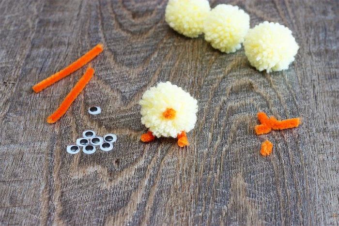 sticking orange fuzzy wire, on pale yellow pom pom, easter crafts for kids, supply of eye stickers , pom poms and fuzzy wire nearby