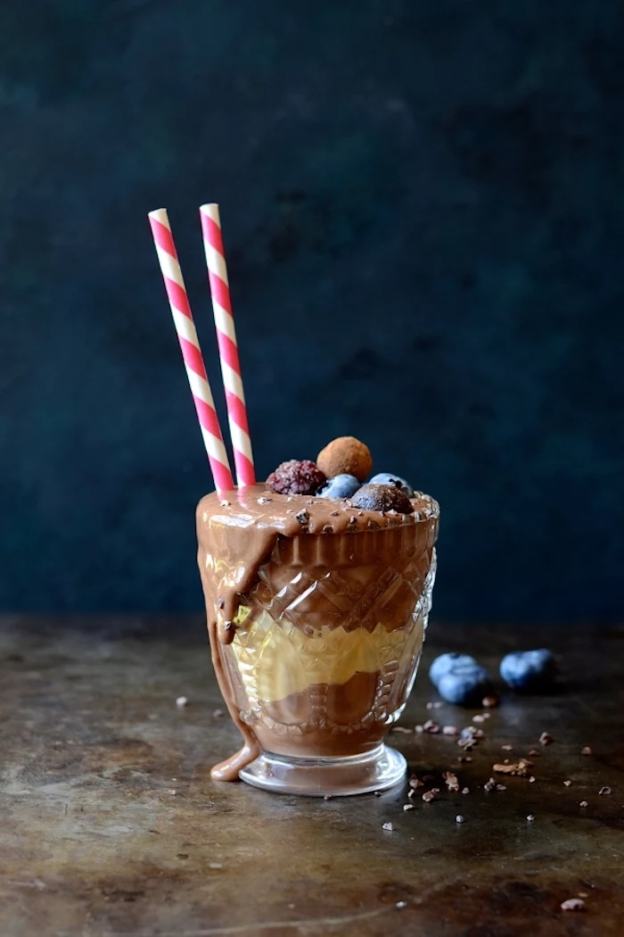 protein shake recipes, small crystal glass, filled with a chocolate smoothie, decorated with blueberries, chocolate chips and two straws