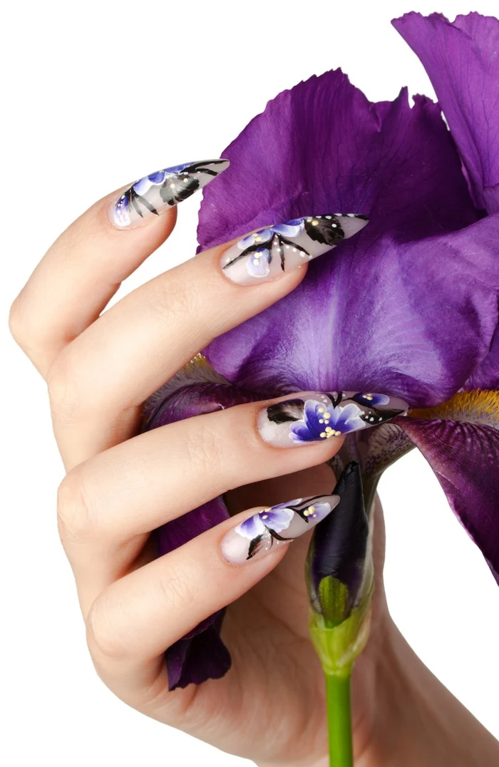 flowers in blue and black, yellow and , painted on long, matte acrylic nails, stilleto nail designs, on hand holding purple flower